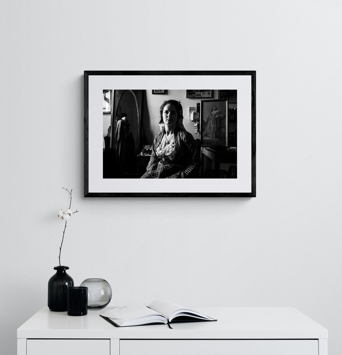 Black and white portrait of a woman in a room with framed pictures on the wall wearing the bridal dress of Soufli, Thrace, Greece. Photography framed on a wall by George Tatakis