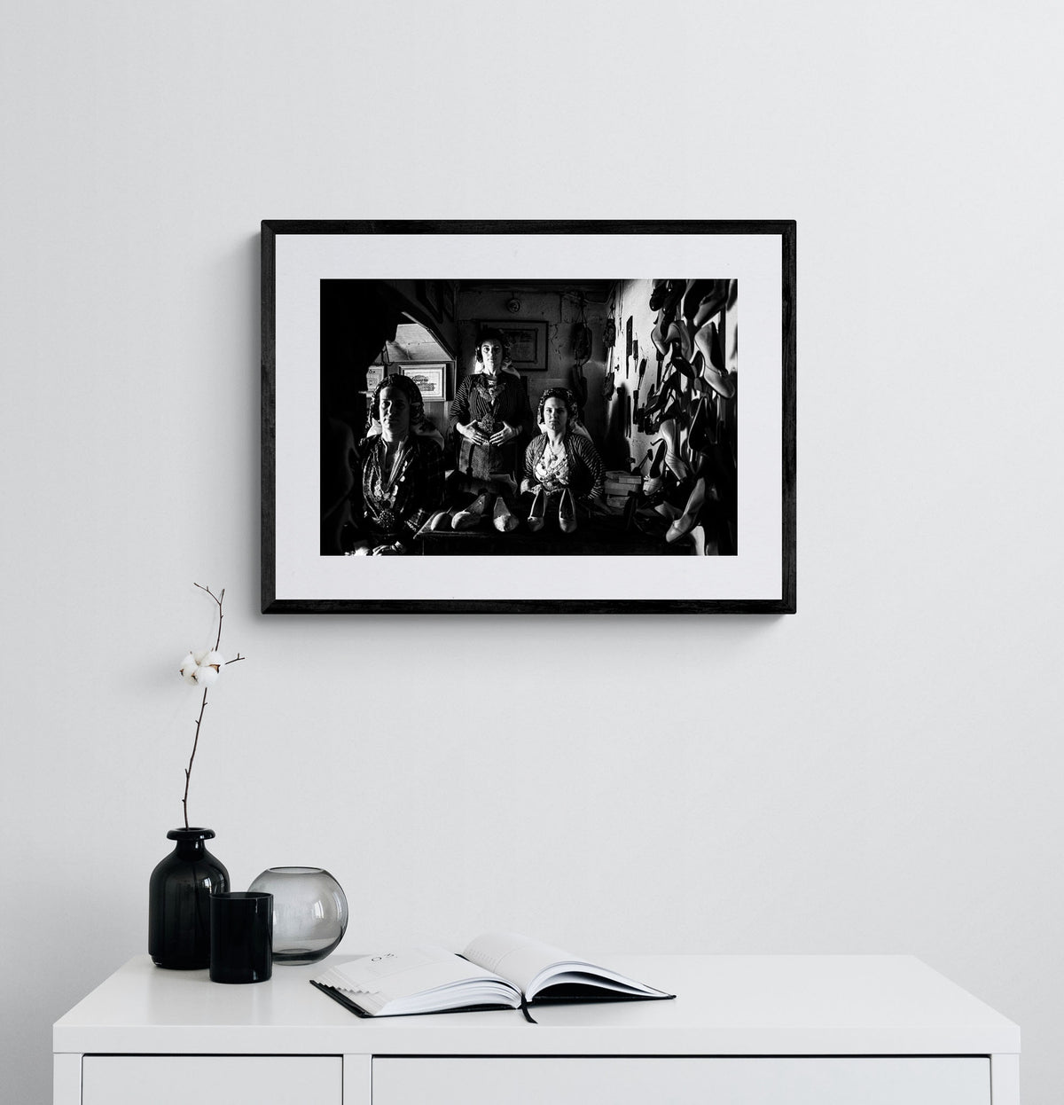 Three women in a room with shelves and shoes. Traditional costumes of Soufli, Thrace, Greece. Black and white photography framed on a wall by George Tatakis