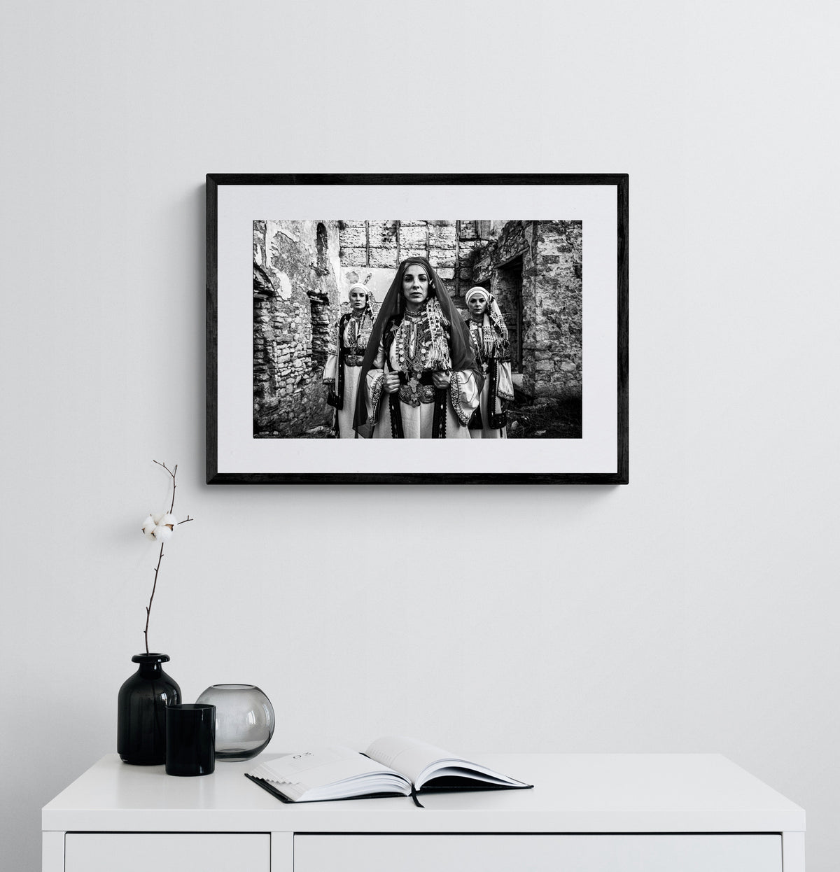 Unveiled bride in traditional attire standing in front of a stone wall. Kyparissia, Peloponnese, Greece. Black and white photography framed on a wall by George Tatakis.